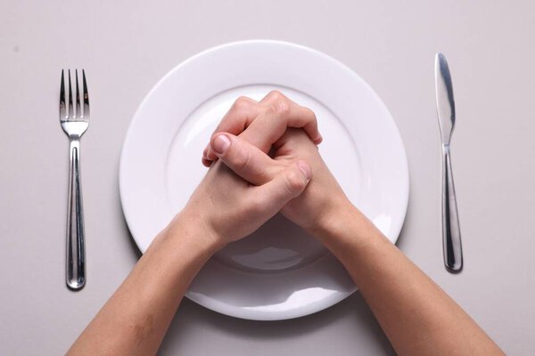 Человек молится перед едой за светло-серым столом с тарелкой и столовыми приборами, вид сверху