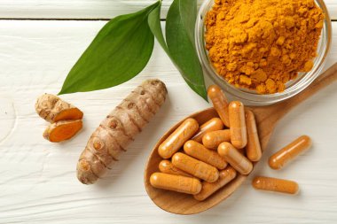 Zerdeçal kapsülleri, toz, kökler ve yeşil yapraklar beyaz ahşap masada, düz yerde.