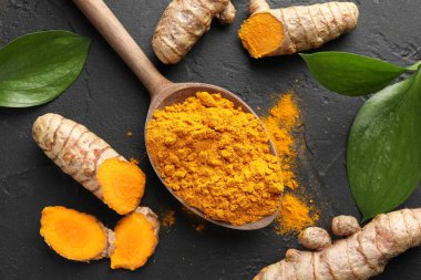 Kaşıkta aromatik zerdeçal tozu, taze kökler ve siyah masa üzerindeki yapraklar, düz uzanmış.