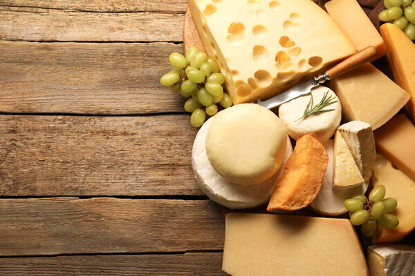 Different types of cheese, rosemary and grapes on wooden table, flat lay. Space for text