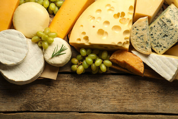Different types of cheese, rosemary and grapes on wooden table, flat lay. Space for text