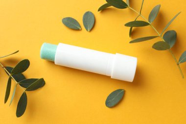 Dudak kremi ve okaliptüs yaprakları sarı arka planda, düz yerde.