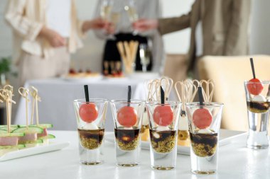 Beyaz masada bir sürü lezzetli kanepe var. İnsanlar kapalı mekanlarda şarap bardaklarını tokuşturuyor.