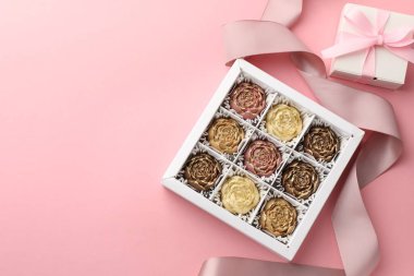 Kutuda çiçek şeklinde çikolata bonbonları, pembe arka planda hediye ve kurdele, düz yatak. Metin için boşluk