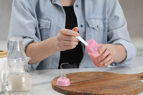 Mother making baby formula in feeding bottle at table indoors, closeup