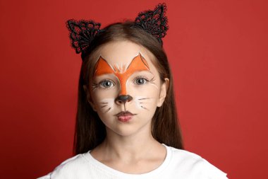 Yüzü boyalı ve kulakları kırmızı arka planda kedi gibi olan tatlı bir kız.