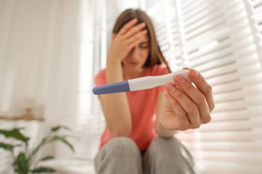 Evdeki pencerenin yanında negatif hamilelik testi olan üzgün kadın, seçici odaklı.