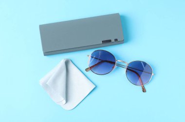 Case with stylish sunglasses and cleaning cloth on light blue background, flat lay