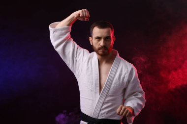 Üniformalı bir adam renkli ışıklarla karate yapıyor ve siyah arka planda sigara içiyor.