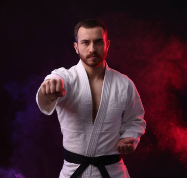 Üniformalı bir adam renkli ışıklarla karate yapıyor ve siyah arka planda sigara içiyor.