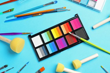 Açık mavi arka planda parlak yüz boyaları, fırçalar ve süngerler, düz yüz.