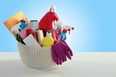 Different cleaning products and supplies in basket on wooden table against light blue gradient background, space for text