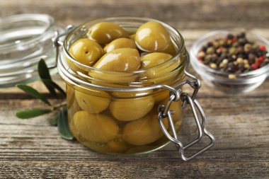 Cam kavanozda turşulu yeşil zeytin ve ahşap masada baharatlar, yakın plan.