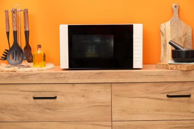 Microwave oven and kitchen utensils on wooden cabinet near orange wall indoors