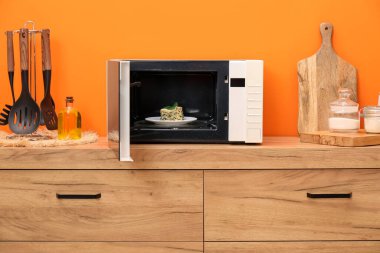 Mikrodalga fırını lazanya ve mutfak gereçleri ile birlikte kapalı alandaki turuncu duvarın yanındaki ahşap dolapta.