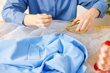 Kadın iç mekândaki mavi gömleğe iğne ve iplikle düğme dikiyor.