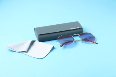 Case with stylish sunglasses and cleaning cloth on light blue background