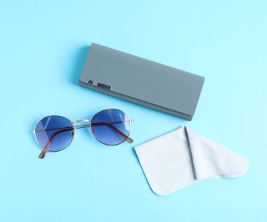 Case with stylish sunglasses and cleaning cloth on light blue background, flat lay