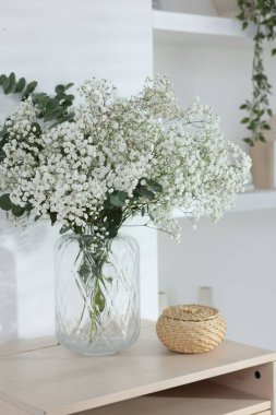 Bouquet of beautiful gypsophila flowers and eucalyptus branches in vase on chest of drawers near white wall