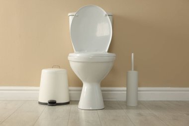 Toilet bowl, brush and trash bin near beige wall in restroom