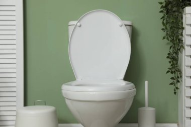 Ceramic toilet bowl, brush and trash can near green wall indoors