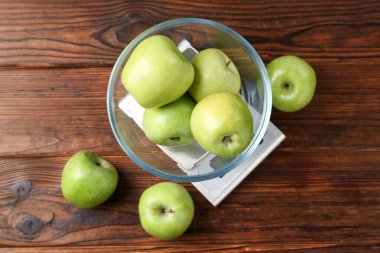 Electronic kitchen scale with bowl of green apples on wooden table, flat lay