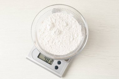 Electronic kitchen scale with bowl of flour on white wooden table, top view