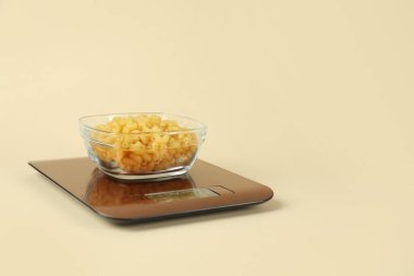 Electronic kitchen scale with bowl of raw pasta on beige background. Space for text