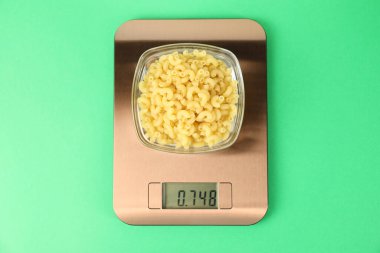 Electronic kitchen scale with bowl of raw pasta on green background, top view