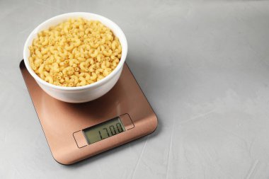 Electronic kitchen scale with bowl of raw pasta on grey textured table. Space for text