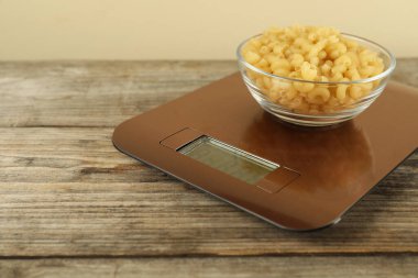 Electronic kitchen scale with bowl of raw pasta on wooden table against beige background, closeup. Space for text