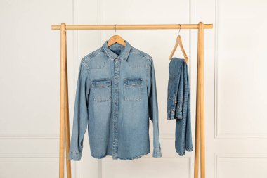 Stylish denim jacket and jeans hanging from rack near white wall