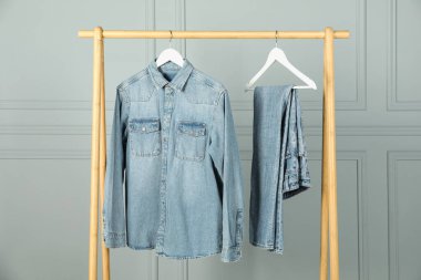 Stylish denim jacket and jeans hanging from rack near white wall