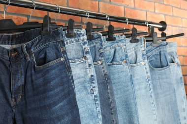 Many stylish jeans hanging from rack near brick wall, closeup