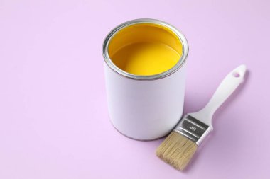 Can of yellow paint and brush on violet background, closeup. Space for text