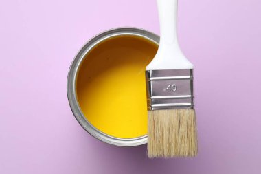 Can of yellow paint and brush on violet background, top view