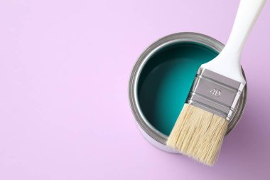 Can of turquoise paint and brush on violet background, top view. Space for text