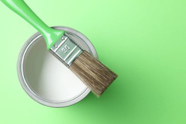 Can of white paint and brush on light green background, top view. Space for text