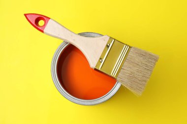 Can of orange paint and brush on yellow background, top view