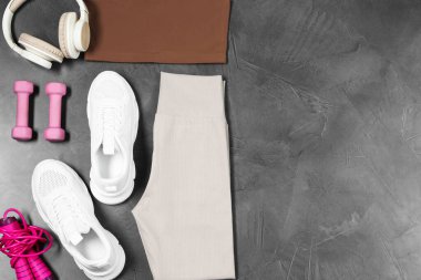 Şık spor kıyafetleri, dambıllar, beyaz spor ayakkabılar, gri arka planda ip atlama ve kulaklıklar, düz yatış. Metin için boşluk