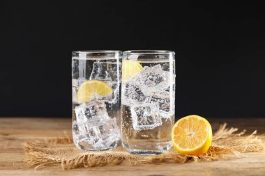 Soda suyunu bardaklarda buzla tazelemek ve ahşap masada limonu siyah arka planda kesmek, yakın plan.