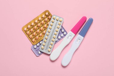 Blisters of oral contraceptive pills and pregnancy tests on pink background, flat lay