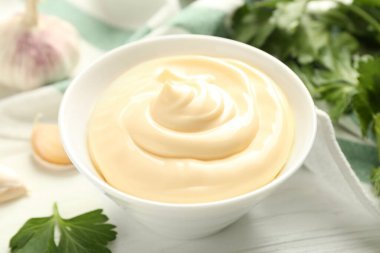 Delicious mayonnaise sauce in bowl, garlic and parsley on white wooden table, closeup
