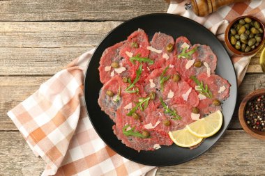 Delicious beef carpaccio with arugula, capers, cheese and lemon on wooden table, flat lay