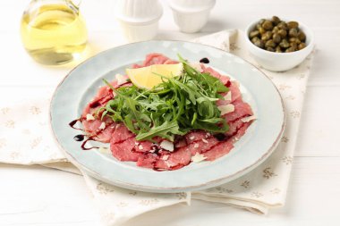 Delicious beef carpaccio with arugula, lemon, capers and oil on white wooden table, closeup