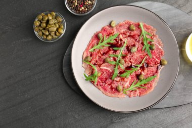 Fresh beef carpaccio with arugula, capers and sauce on dark textured table, flat lay. Space for text