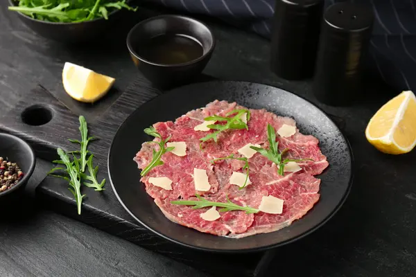 Fresh beef carpaccio with arugula and cheese on dark textured table, closeup