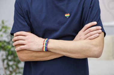 Kalp şeklinde iğnesi ve bileziği olan bir adam. Kapalı alanda LGBT bayrağı renginde, yakın plan.