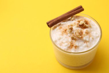 Tasty rice pudding with cinnamon and nuts in glass bowl on yellow background, closeup. Space for text
