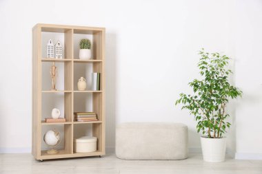 Shelving unit with different home decor, houseplant and ottoman near white wall indoors